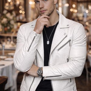 Peut inclure: Veste en cuir blanc texturé avec des fermetures éclair argentées, portée sur un t-shirt noir. La veste est de style motard. Une montre et un collier argentés complètent le look. L'arrière-plan est un restaurant flou.