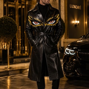 May include: A black trench coat featuring a graphic print of two stylized yellow and orange eyes on the chest area. The coat has a wide collar, long sleeves, a belted waist, and a smooth finish. It is worn over a black turtleneck and black trousers.