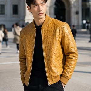 May include: A mustard yellow bomber jacket featuring a textured ostrich-skin pattern, a ribbed collar, ribbed cuffs, and a central silver zipper closure. The jacket is worn over a plain black shirt and paired with black trousers.