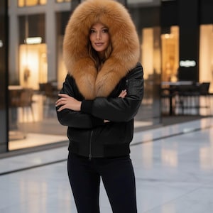 Peut inclure: Veste de bombardier noire avec une grande capuche bordée de fourrure beige. La veste a une fermeture éclair et un ourlet côtelé. La personne porte un pantalon bleu foncé et des bottes marron. L'arrière-plan est une zone commerciale intérieure floue.