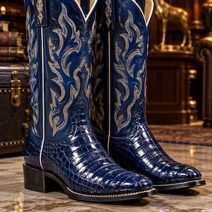 May include: A pair of deep blue cowboy boots featuring a glossy, textured crocodile-skin pattern on the foot and a tall shaft with intricate, light-colored flame-like embroidery. The boots have white piping along the seams and a stacked heel.