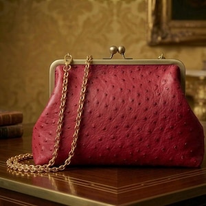 Könnte beinhalten: Eine rote Leder-Clutch mit goldener Kette. Die Tasche hat einen Kiss-Lock-Verschluss und eine strukturierte Oberfläche. Die Kette liegt über der Vorderseite. Die Tasche steht auf einer Holzoberfläche.