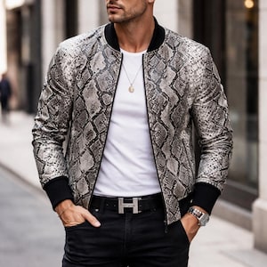 Puede incluir: Chaqueta con estampado de piel de serpiente con ribetes negros, sobre camiseta blanca y pantalón negro. La chaqueta tiene cremallera y puños de canalé. Accesorios: hebilla plateada, reloj y gafas de sol.