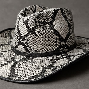 May include: Cowboy hat with a snakeskin pattern in black and white. The hat has a wide brim and a structured crown, with a black band around the base of the crown. The hat is made of a textured material.
