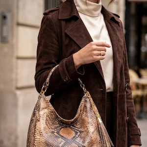 May include: A brown leather shoulder bag with a snakeskin pattern, gold chain straps, and a tassel. The bag is held by a person wearing a brown suede coat and a cream-colored turtleneck.
