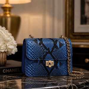 May include: A blue handbag with a snakeskin pattern and a gold chain strap. The bag has a rectangular shape with a gold clasp closure. The word "CHANEL" is visible on a book in the background.