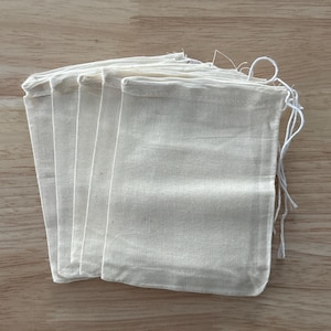 May include: A stack of small, off-white muslin drawstring bags. The bags are rectangular with a rounded top and a white drawstring closure. The bags are made of a light, textured fabric and are arranged on a light brown wooden surface.
