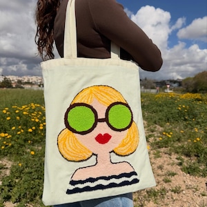 Op de afbeelding: Crèmekleurige draagtas met een vrouwengezicht. Het gezicht heeft geel haar, groene zonnebril en rode lippen. De tas heeft een zwart-wit gestreepte bovenkant en crèmekleurige handvatten. De tas wordt buiten gedragen in een veld met gele bloemen.