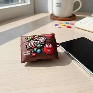 Puede incluir: Una funda marrón para AirPods con forma de envoltorio de barra de caramelo, con el logotipo de M&M's y caramelos de colores. La funda tiene un personaje de M&M's rojo en la parte delantera. Un smartphone está al lado de la funda.