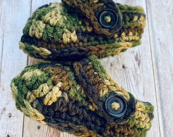 Könnte beinhalten: Ein Paar gehäkelte Babyschuhe in Tarnmuster mit schwarzen Knöpfen. Die Schuhe sind aus grünem, braunem und beigefarbenem Garn gefertigt.
