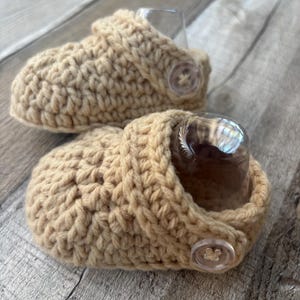 May include: A pair of beige crochet baby booties. Each bootie features a button closure and a thick, textured stitch. The booties are designed to fit small feet and are likely handmade.