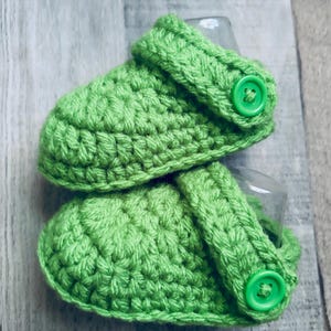 May include: A pair of bright green crochet baby shoes. The shoes have a textured stitch pattern and a strap with a green button closure. The shoes are designed to look like clogs.