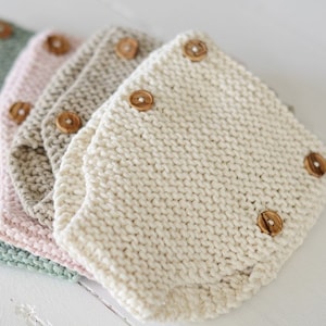 May include: A set of four knitted baby bloomers in different colours, including sage green, pink, beige, and white. Each bloomer has wooden buttons.