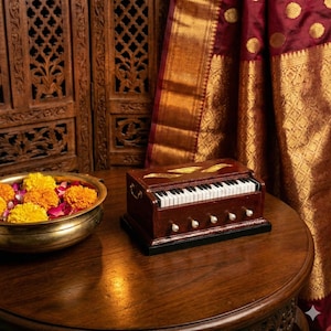 Könnte beinhalten: Ein Miniatur-Harmonium aus Holz mit einer Tastatur und vier Knöpfen steht auf einem runden Tisch aus dunklem Holz. Das Harmonium ist braun mit goldenen Akzenten. Eine Messingschale mit Blumen und ein bordeauxfarbener und goldener Stoff befinden sich im Hintergrund.
