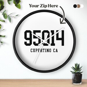 May include: A round wall clock with a black frame and a white face. The clock face displays the numbers "95014" in bold black font, with "CUPERTINO CA" below. The text "Your Zip Here" is above the clock.
