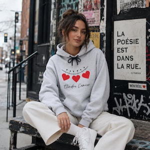 Peut inclure: Un sweat à capuche gris clair avec un cordon de serrage noir et un graphisme de trois cœurs rouges reliés. Le texte "Étendre l'amour" est en dessous des cœurs. La personne porte un pantalon blanc et des baskets.