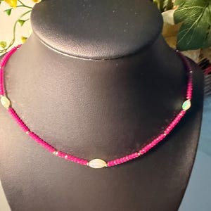 Peut inclure: Un collier de perles composé de petites perles facettées rose vif alternées avec trois pierres blanches ovales, lisses et irisées. Le collier est assemblé avec de minuscules perles intercalaires dorées entre les éléments roses et blancs.