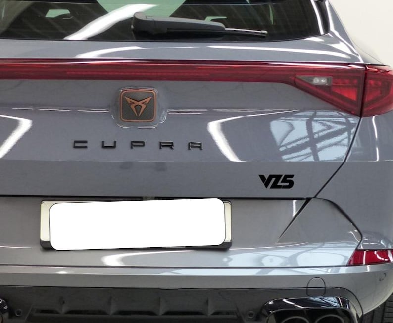 May include: Rear view of a gray SUV with the word "CUPRA" in black lettering. A bronze emblem is above the lettering. The model designation "VZ5" is in black on the right side of the vehicle.