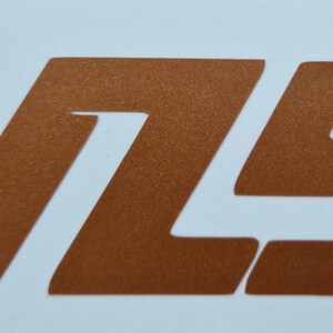 May include: A copper-colored graphic of the letters "V75" on a white background. The numbers and letter are in a bold, sans-serif font. The image is a close-up, with the letters and numbers taking up most of the frame.