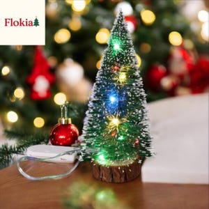 Peut inclure: Un petit sapin de Noël artificiel avec des aiguilles vertes et des lumières LED multicolores. L'arbre est posé sur une base en bois. Une boule rouge et un bloc-piles blanc sont visibles. La marque "Flokia" est dans le coin supérieur gauche.