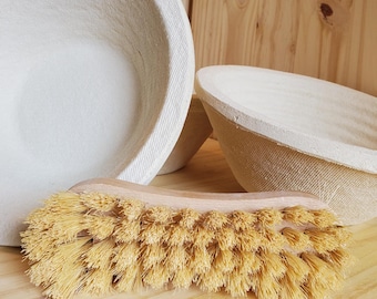 May include: Two round, off-white proofing baskets with a textured, woven surface and a wooden-handled brush featuring stiff, light brown bristles. The baskets have a deep, bowl-like shape, and the brush is positioned horizontally in the foreground.