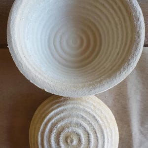 May include: Two round, light-colored proofing baskets with concentric circular patterns. The baskets are made of a textured material, likely natural fibers, and are designed for bread baking. One basket is inverted, revealing its interior.