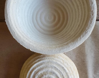 May include: Two round, light-colored proofing baskets with concentric circular patterns. The baskets are made of a textured material, likely natural fibers, and are designed for bread baking. One basket is inverted, revealing its interior.