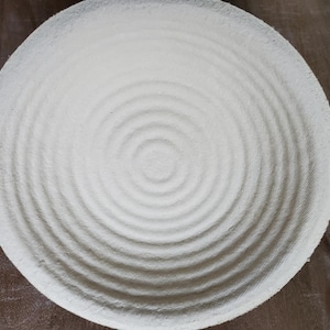 May include: A round, off-white woven bowl with a concentric spiral design. The bowl's texture is visible, with a series of raised, circular ridges radiating from the center. The bowl is likely used for serving or display.