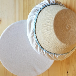 May include: Two round, beige objects on a light wood surface. One is a woven basket with a white fabric cover and blue stitching. The other is a flat, white, circular object with a ribbed texture.
