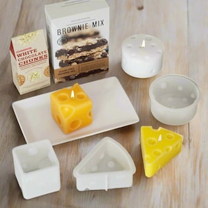 Pode incluir: Uma coleção de velas de novidade e ingredientes para assar. As velas têm a forma de queijo e têm buracos. Há uma caixa de mistura para brownie e um saco de pedaços de chocolate branco. As velas estão em um prato branco.