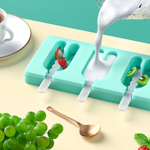 Może przedstawiać: Jasnoturkusowa forma do lodów na patyku z białymi patyczkami, wypełniona truskawkami, kiwi i wlewanym białym płynem. Złota łyżka i zielone winogrona znajdują się na pierwszym planie. Biała filiżanka i spodek są w tle.