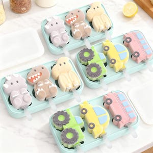 Może przedstawiać: Kolorowe foremki do lodów w kształcie dinozaurów, traktorów, samochodów i autobusów. Formy są ułożone w jasnoniebieskich tackach, każda z patyczkiem. Lody są w różnych kolorach, idealne na lato.