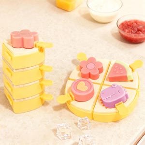 Può includere: Stampi per ghiaccioli gialli e rosa. Una pila di quattro stampi con cime a forma di fiore e manici gialli. Un secondo set di stampi è diviso in quattro sezioni, ciascuna con una forma diversa: fiore, cuore, anguria e dinosauro.