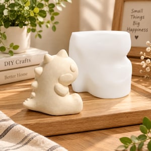 Può includere: Una statuetta a forma di dinosauro color beige con piccole punte lungo la schiena e una finitura liscia e opaca, posizionata accanto a uno stampo in silicone bianco, arrotondato e semplice su una superficie in legno.