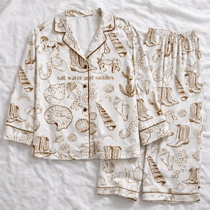 May include: White pajama set with a brown pattern of cowboy boots, seashells, horses, and cacti. The set includes a button-down top and matching pants. The text "salt water and saddles" is printed on the top.