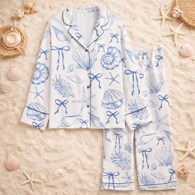 May include: White pajama set with a blue seashell and bow pattern. The long-sleeved top has a button-down front and navy trim. The matching pants have an elastic waistband. The set is designed for comfort and relaxation.