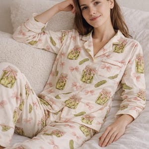 May include: Cream-coloured pyjama set with a repeating pattern of pickle jars and pink bows. The long-sleeved top has a button-down front and a chest pocket. The matching trousers complete the comfortable sleepwear ensemble.
