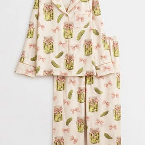 May include: Cream-coloured pyjama set with a repeating pattern of pickle jars tied with pink bows and individual pickles. The long-sleeved top has a collared neckline and button closure. The set is trimmed with light brown piping.