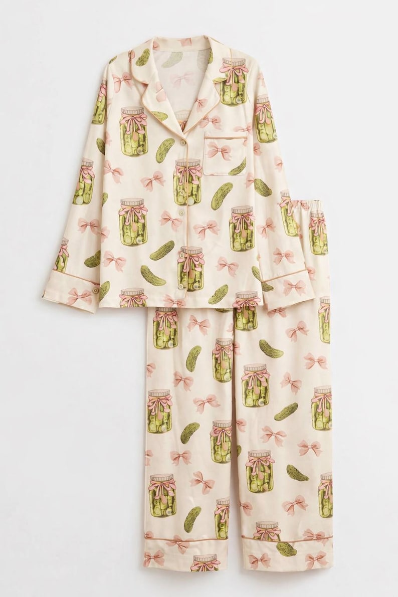 May include: Cream-coloured pyjama set with a repeating pattern of pickle jars tied with pink bows and individual pickles. The long-sleeved top has a collared neckline and button closure. The set is trimmed with light brown piping.