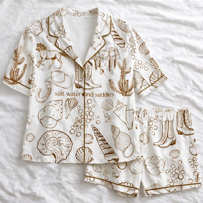 May include: White pajama set with a brown illustration of seashells, cowboy boots, a horse, and cacti. The set includes a short-sleeved button-up shirt and matching shorts. The text "salt water and saddles" is printed on the shirt.