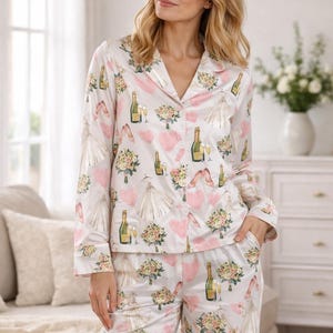 May include: White satin pajama set with a repeating pattern of pink hearts, wedding dresses, champagne bottles, bouquets, and high heels. The long-sleeved top has a notched collar and button-down front. The pants have a relaxed fit.