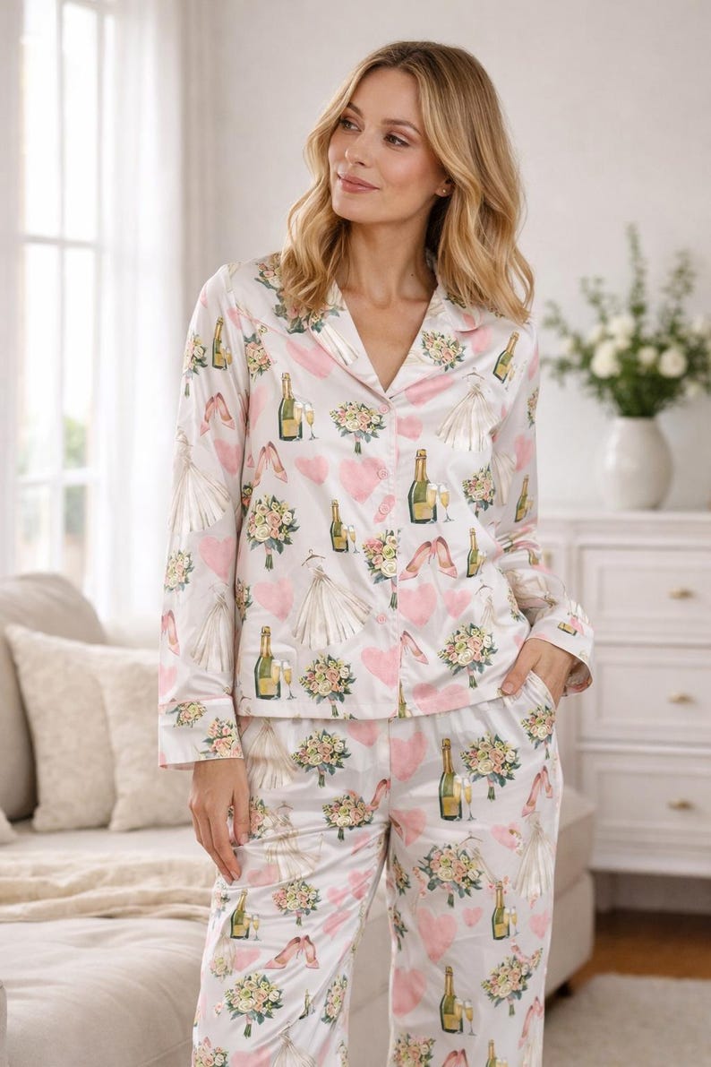 May include: White satin pajama set with a repeating pattern of pink hearts, wedding dresses, champagne bottles, bouquets, and high heels. The long-sleeved top has a notched collar and button-down front. The pants have a relaxed fit.