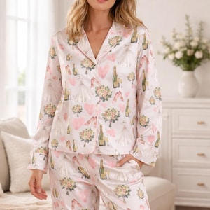 May include: Light pink pajama set with a repeating pattern of wedding-themed illustrations. The pattern includes champagne bottles, wedding dresses, bouquets, and hearts. The set includes a button-down shirt and matching pants.