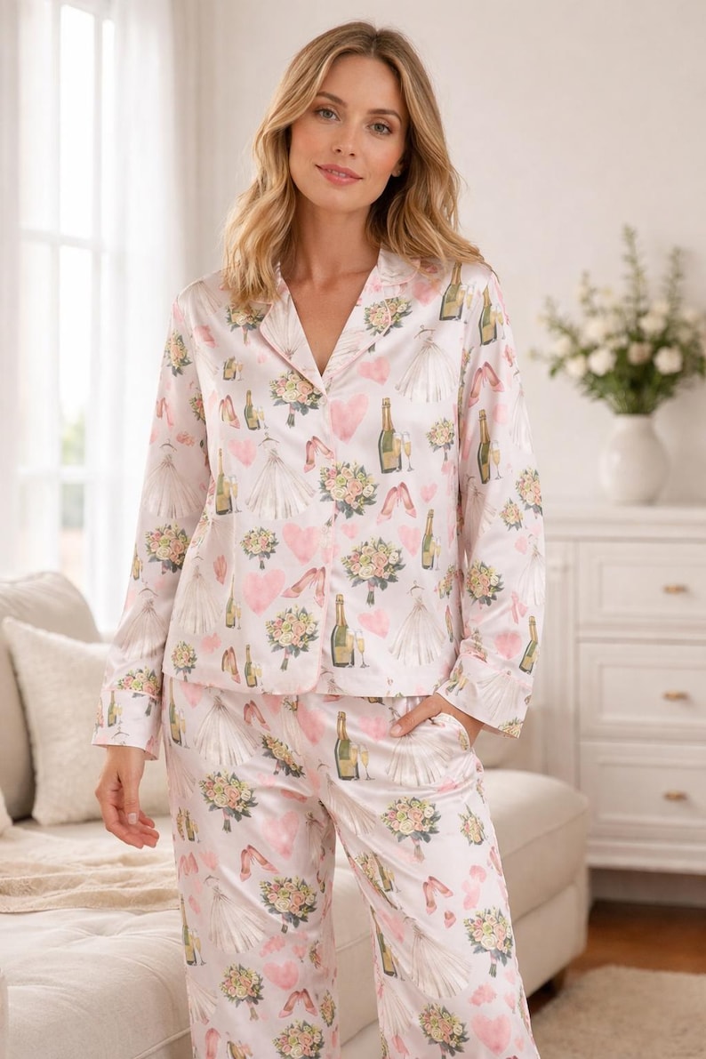 May include: Light pink pajama set with a repeating pattern of wedding-themed illustrations. The pattern includes champagne bottles, wedding dresses, bouquets, and hearts. The set includes a button-down shirt and matching pants.