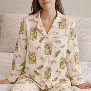 May include: Cream-coloured pyjama set with a repeating pattern of pickle jars and pink bows. The long-sleeved top and trousers have light brown trim. The pyjamas are made of a soft, comfortable material.