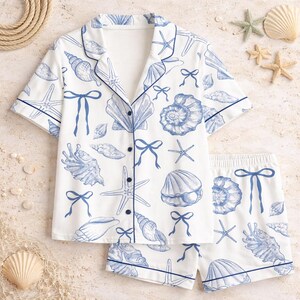 May include: White pajama set with a blue seashell and starfish pattern. The short-sleeved top and shorts have blue trim and button details. The nautical-themed sleepwear set includes a collared shirt and shorts.