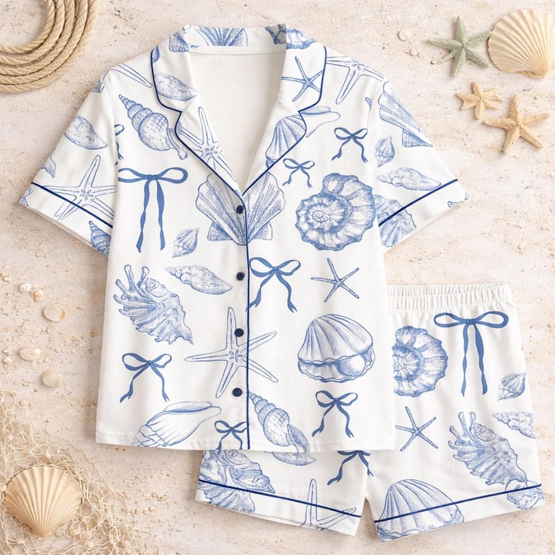 May include: White pajama set with a blue seashell and starfish pattern. The short-sleeved top and shorts have blue trim and button details. The nautical-themed sleepwear set includes a collared shirt and shorts.