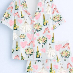 May include: Light blue pajama set with a repeating pattern of champagne bottles, bouquets, high heels, wedding dresses, and pink hearts. The set includes a button-down shirt and matching shorts, trimmed with pink piping.