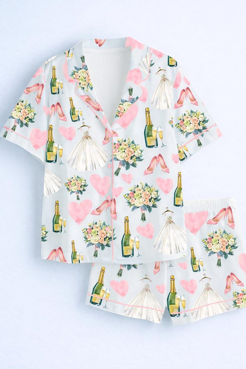 May include: Light blue pajama set with a repeating pattern of champagne bottles, bouquets, high heels, wedding dresses, and pink hearts. The set includes a button-down shirt and matching shorts, trimmed with pink piping.