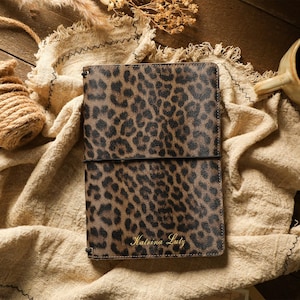 May include: A rectangular notebook cover featuring a brown and black leopard print pattern. The cover is secured with a thin black elastic band and has the name Katrina Lutz embossed in gold lettering at the bottom center of the front.